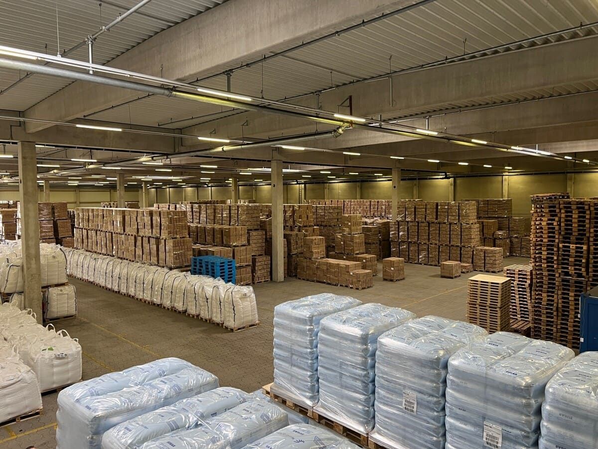 A spacious warehouse filled with stacked pallets of boxes and sacks arranged in neat rows under bright skylights.