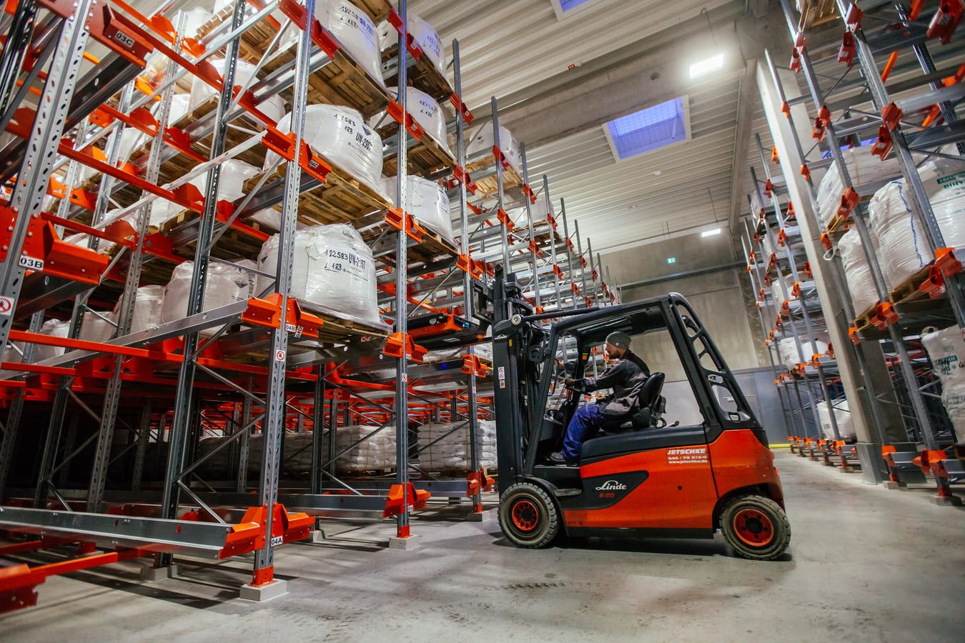 A forklift driver navigates through a spacious warehouse with high shelves stacked with large sacks. The bright lighting improves visibility.