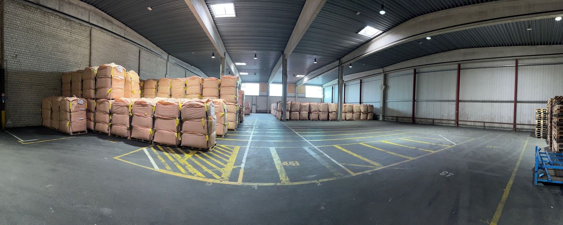 The inside of a spacious warehouse, filled with large, stacked bales secured with yellow straps, next to marked parking lines.